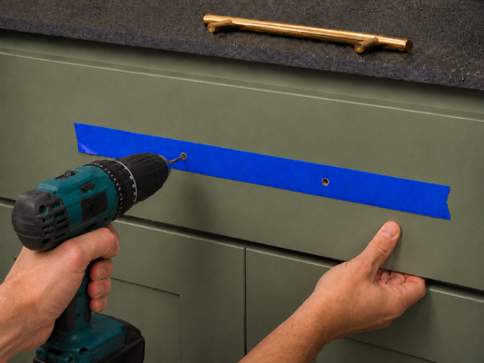 Drilling cabinet pull holes through painter’s tape on a green drawer front before installing the hardware.
