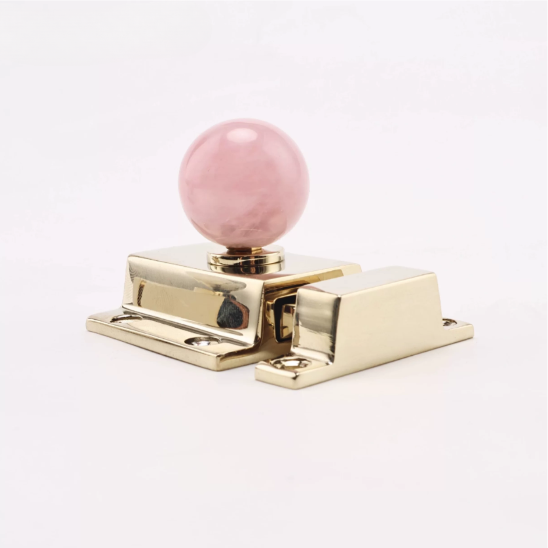 A polished gold-finished metal latch or cabinet hardware piece with a round pink stone knob on top, photographed against a clean white background.