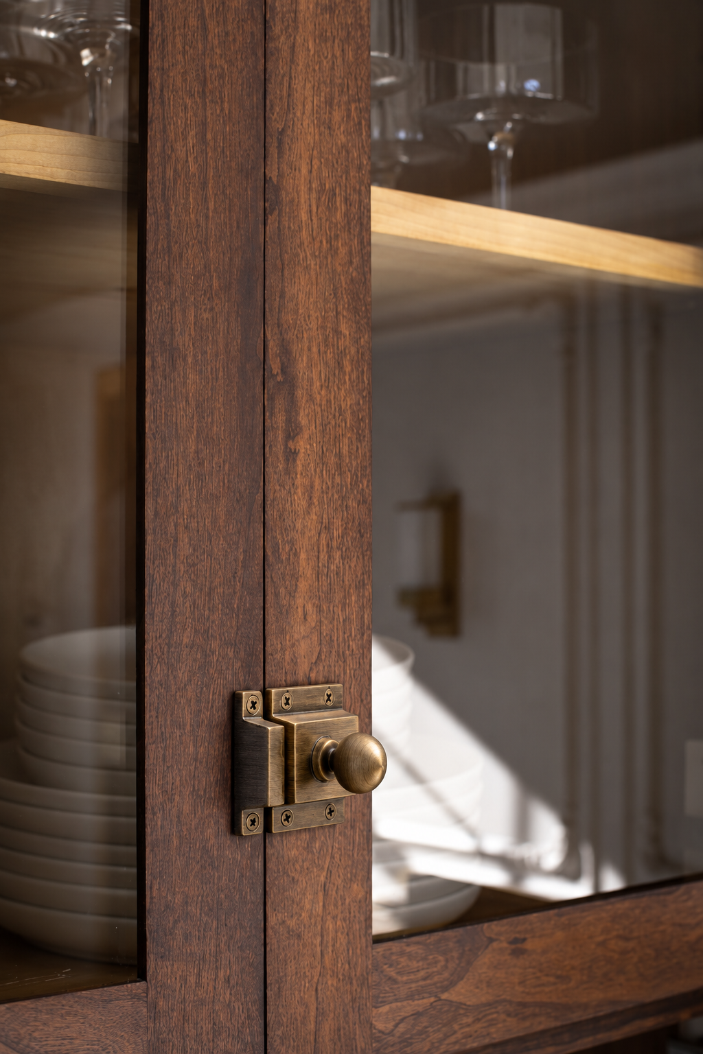 Close-up of a dark brown wooden hutch or cabinet door with a brass latch and round brass knob, showing glass panels and dishes inside.